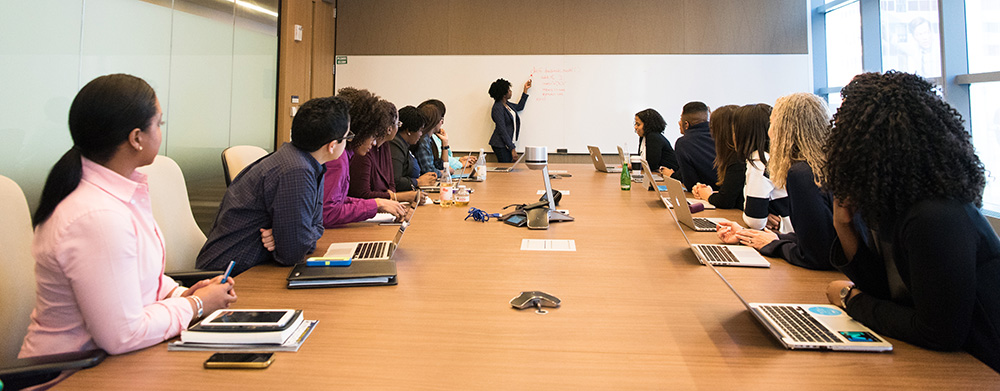 people seated at table in meeting room watching someone speaking at white board